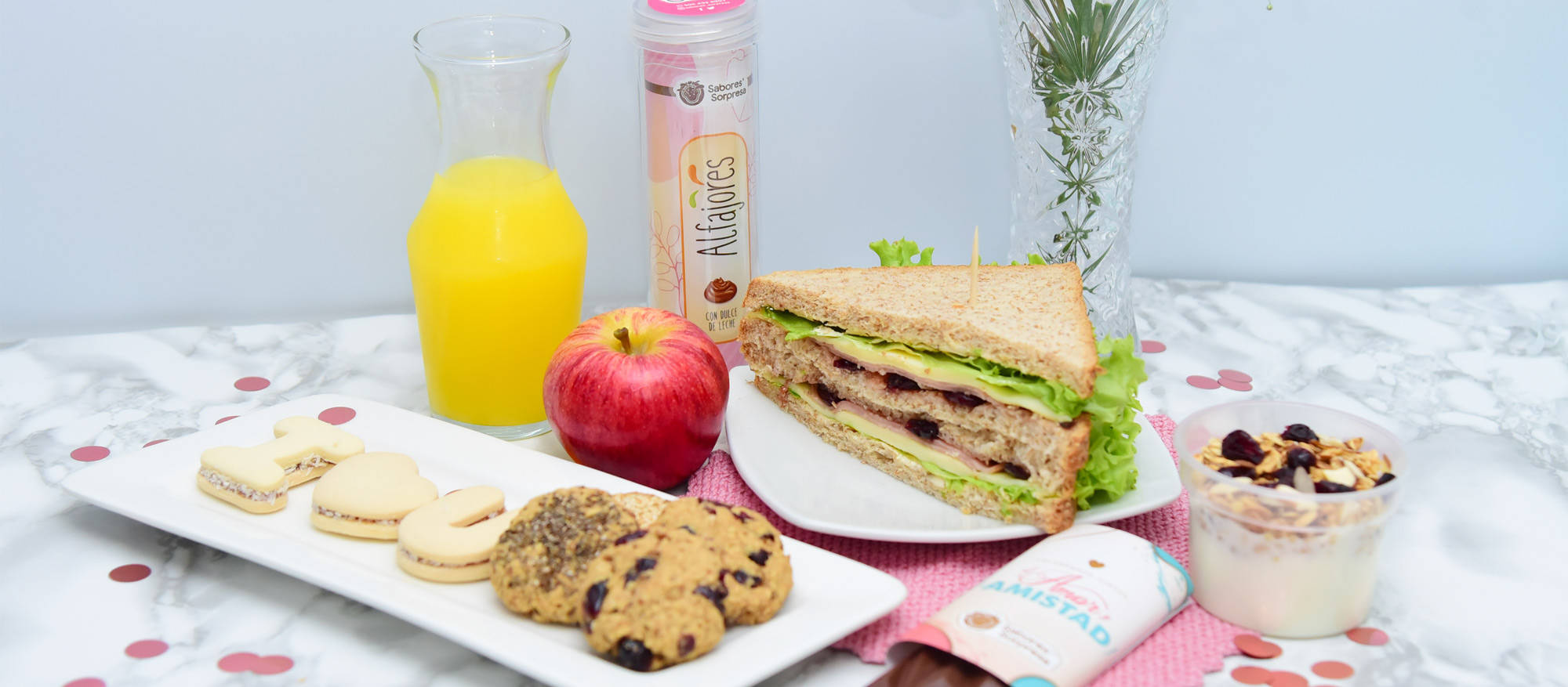 Sánduche triangular con lechuga, queso y jamón, servido en un plato blanco. Al frente unas galletas con forma de la letra I, una forma de corazón, y de la letra U, y algunas galletas redondas de chips de chocolate. Un jarro con jugo de naranja, una manzana, una barra de chocolate y un tazón con yogurt y cereales. Atrás un contenedor de alfajores y un jarro con una rama ornamental.