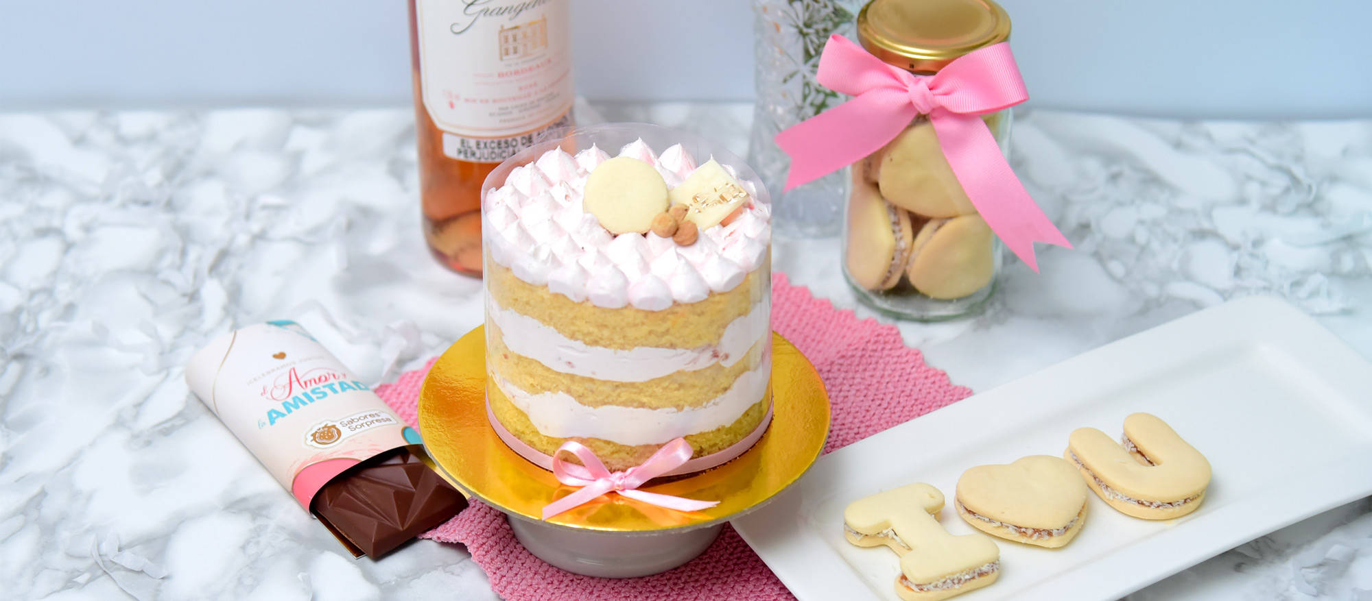 Torta de Vainilla en un plato dorado, a su derecha una barra de chocolate, detrás una botella de vino rosado, a su izquierda unas galletas con las letra I, una forma de corazón, y la letra U y un tarro de galletas con una cinta rosa