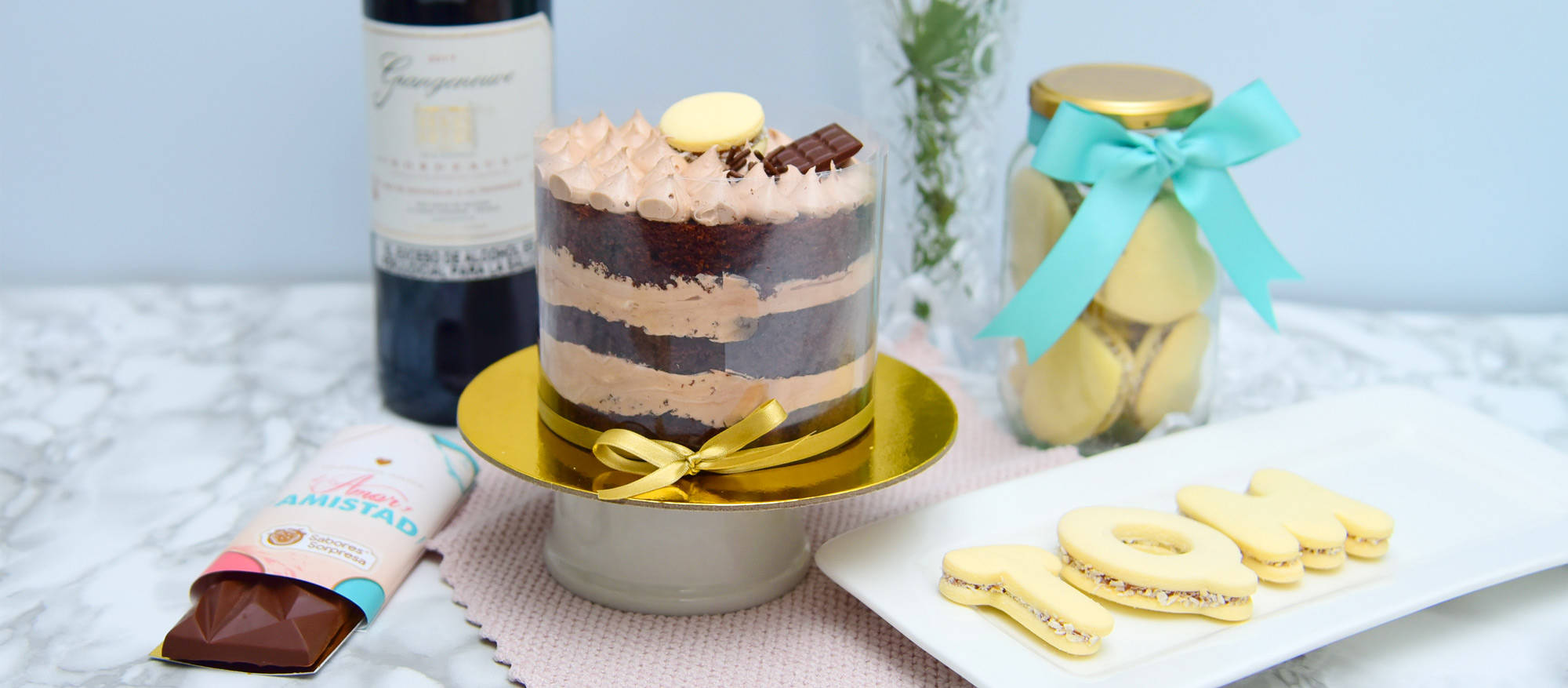 Torta de Chocolate en un plato dorado, a su derecha una barra de chocolate, detrás una botella de vino, a su izquierda unas galletas con las letras TQM y un tarro de galletas con una cinta azul